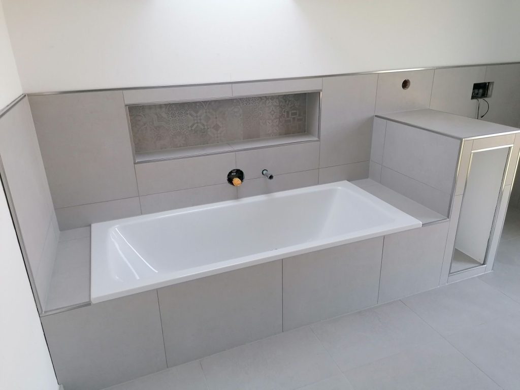 Einbaubadewanne: Weiße Badewanne, bündig in graue Fliesen eingelassen.