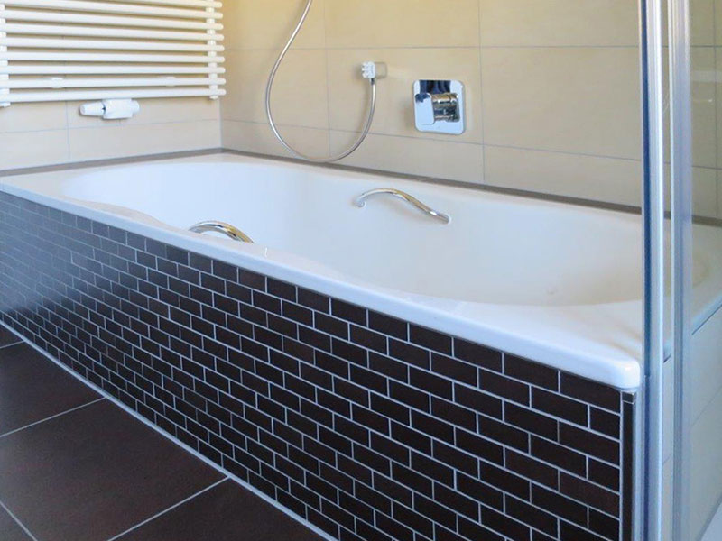 Badewanne: Weiße Einbaubadewanne mit dunkelbrauner Mosaikverkleidung.